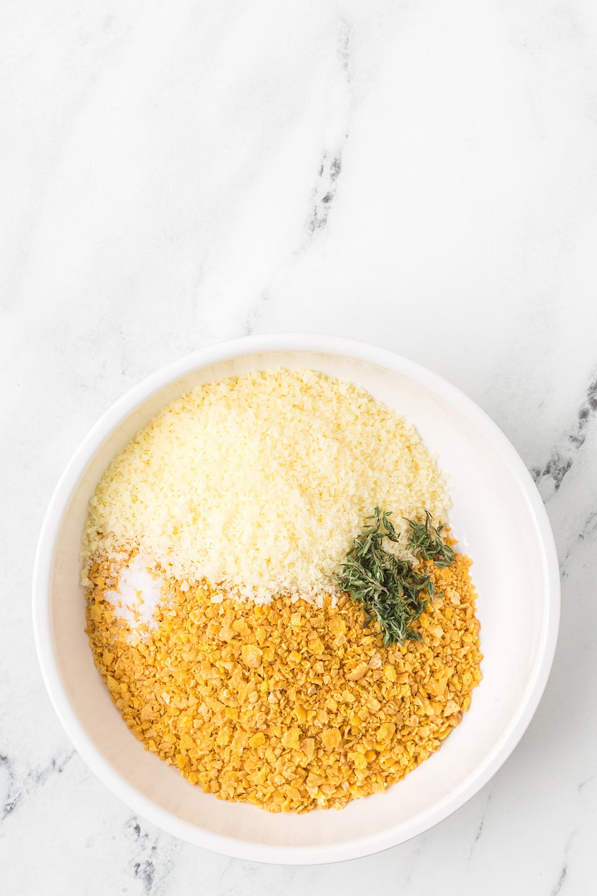White bowl with grated cheese, crushed cornflakes, and fresh herbs on a white marble surface.