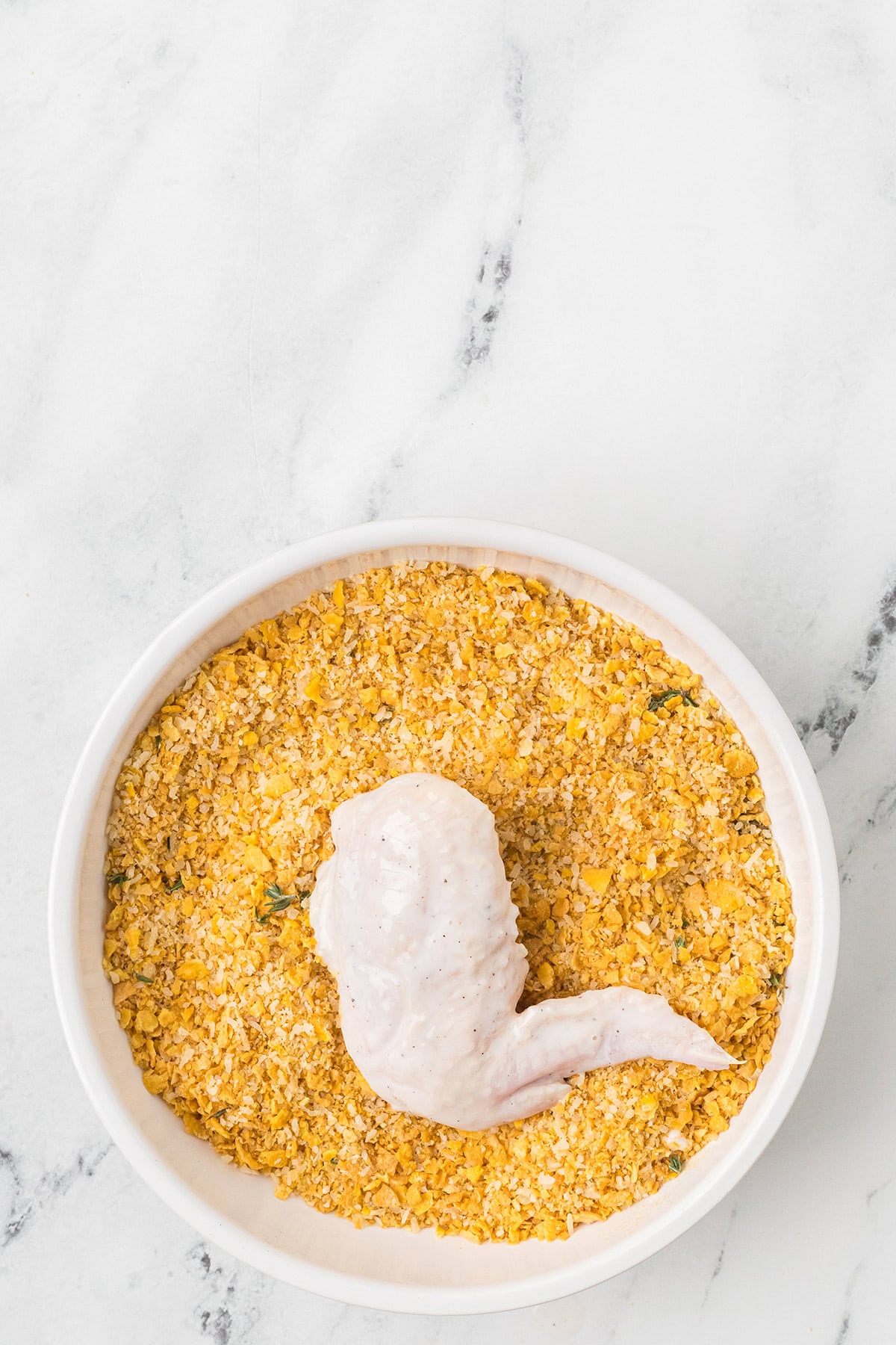 A raw chicken wing coated with marinade sits in a bowl of crushed cornflakes on a marble surface.