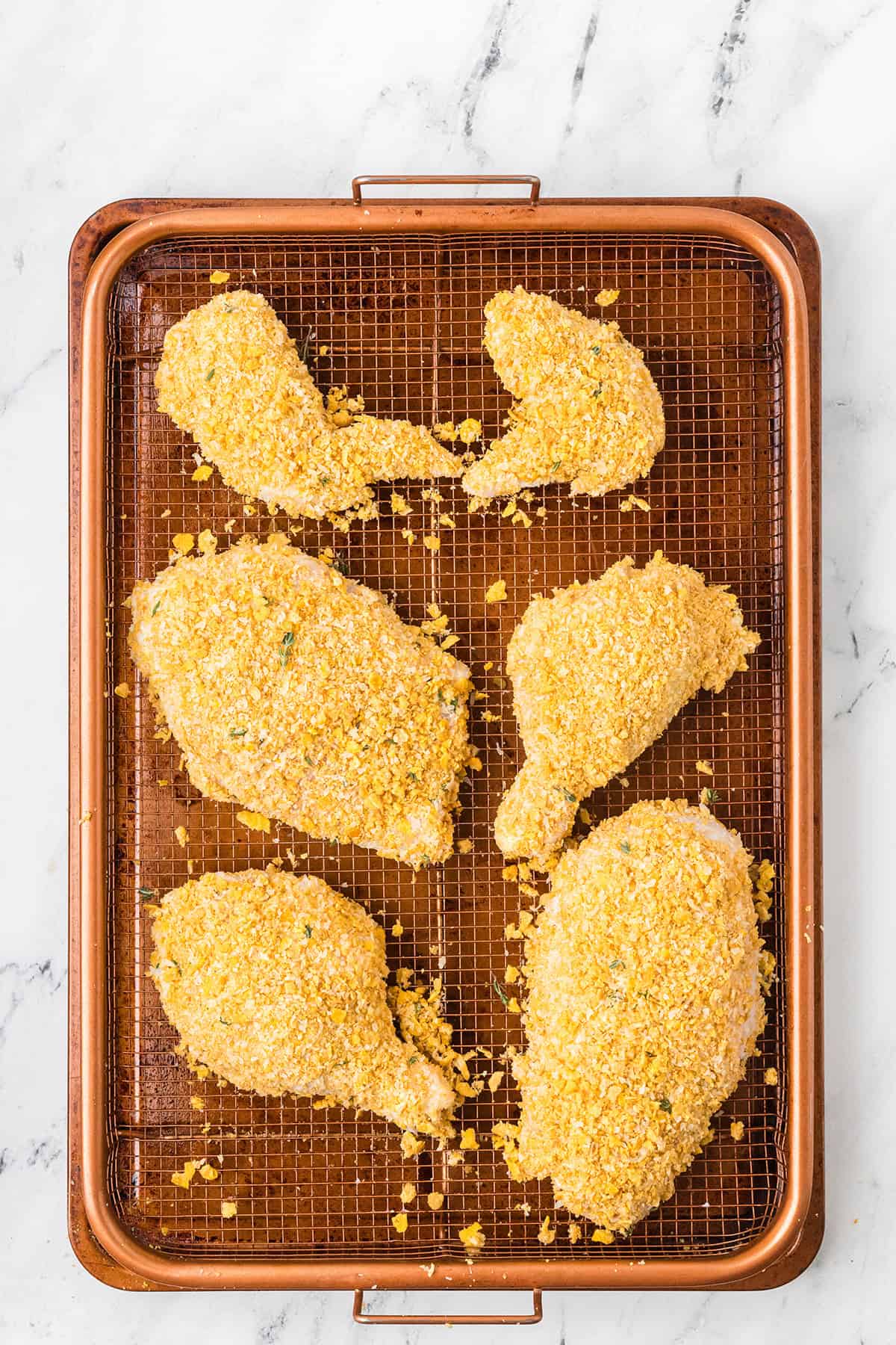 Six breaded chicken pieces on a wire rack set inside a baking tray on a marble surface.