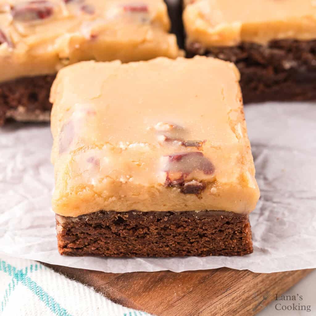A frosted brownie with a thick layer of caramel-colored icing and pecans on parchment paper.