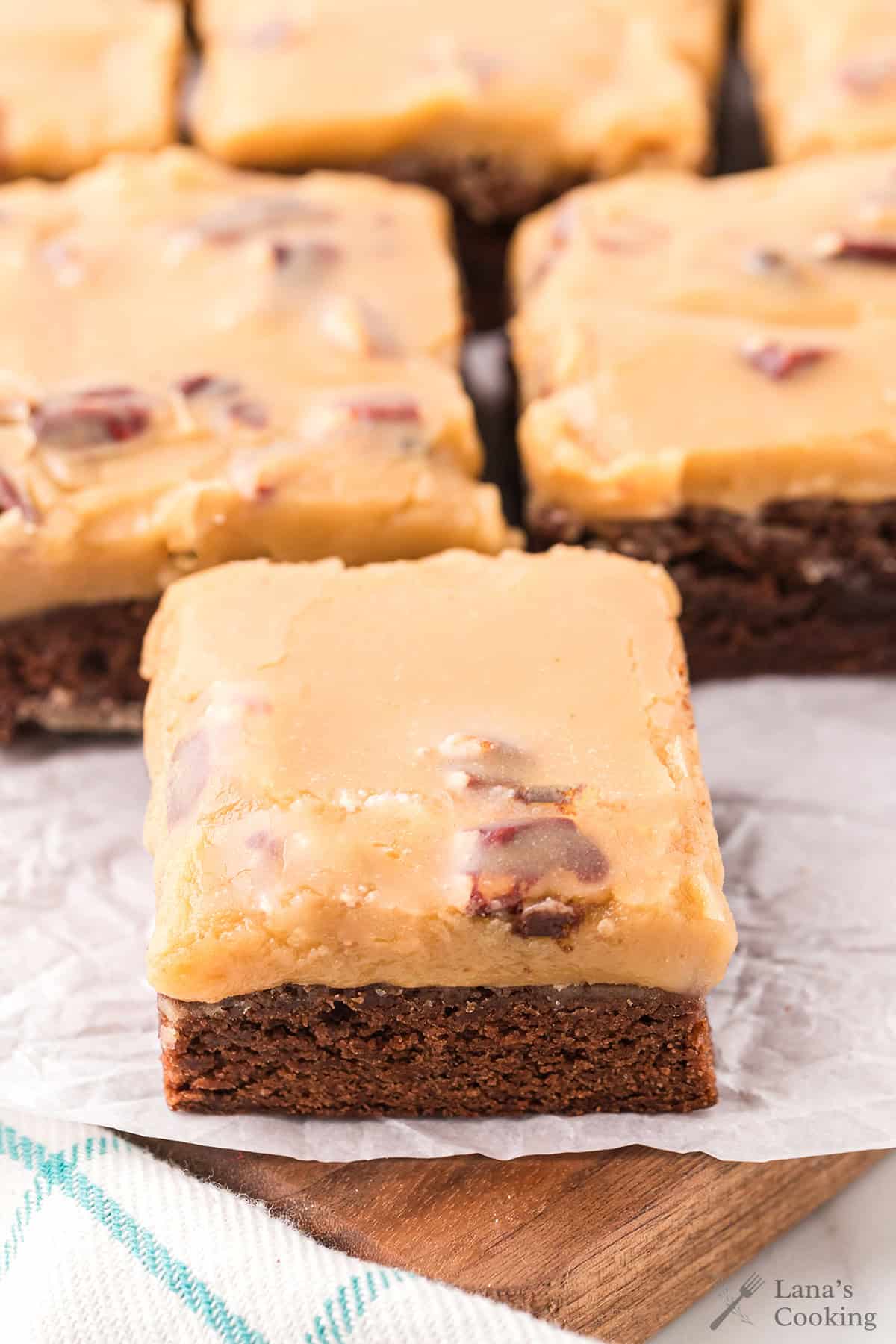 A frosted brownie topped with praline, sitting on parchment paper with more brownies in the background.