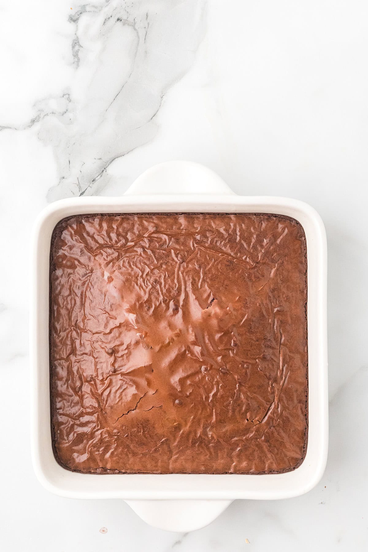 A pan of baked brownies with a shiny, crackly top, in a white square baking dish on a marble surface.