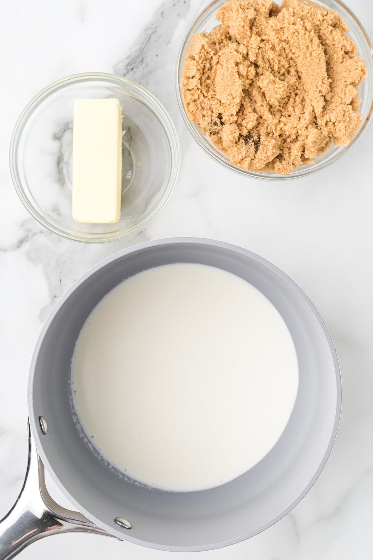 A saucepan of cream, a bowl of brown sugar, and a dish with a stick of butter on a marble surface.