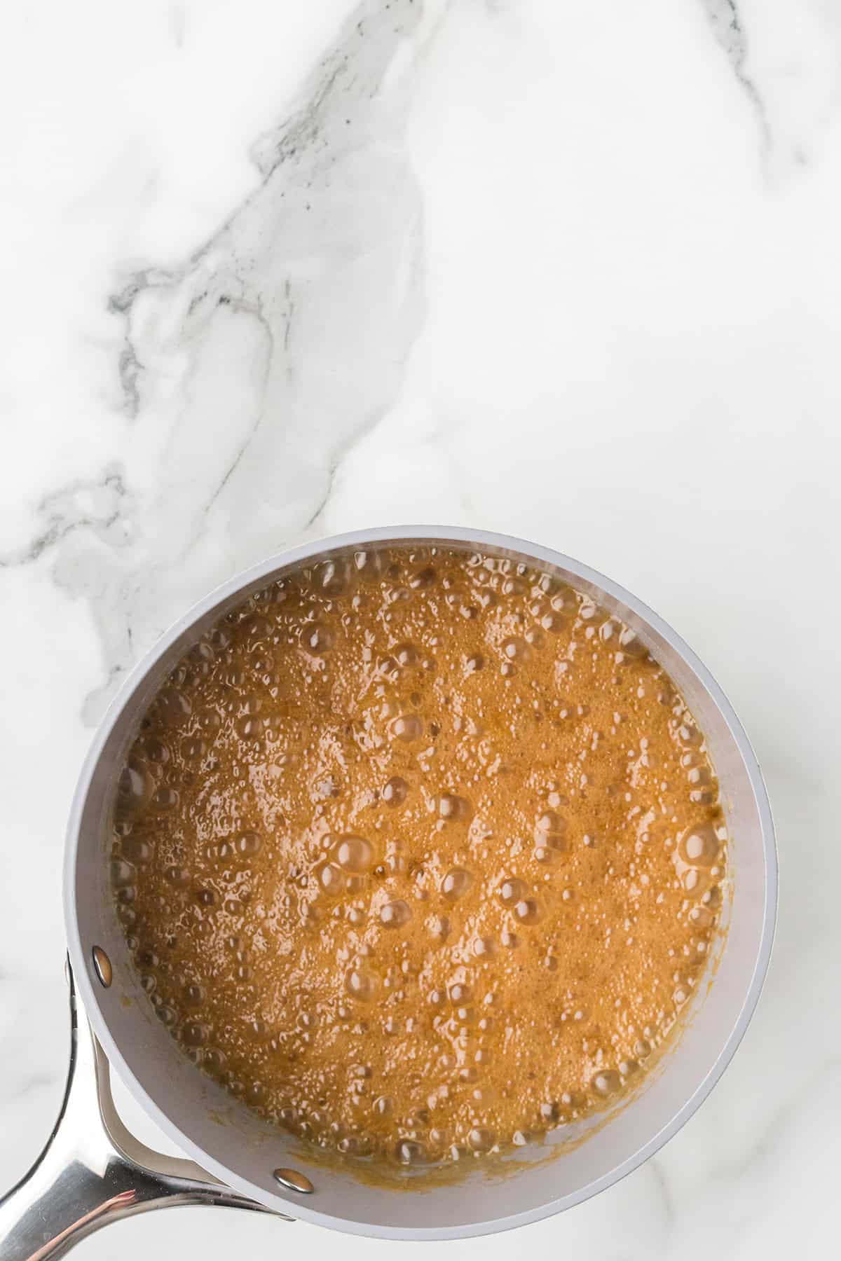 A saucepan with bubbling caramel sauce on a white marble countertop.