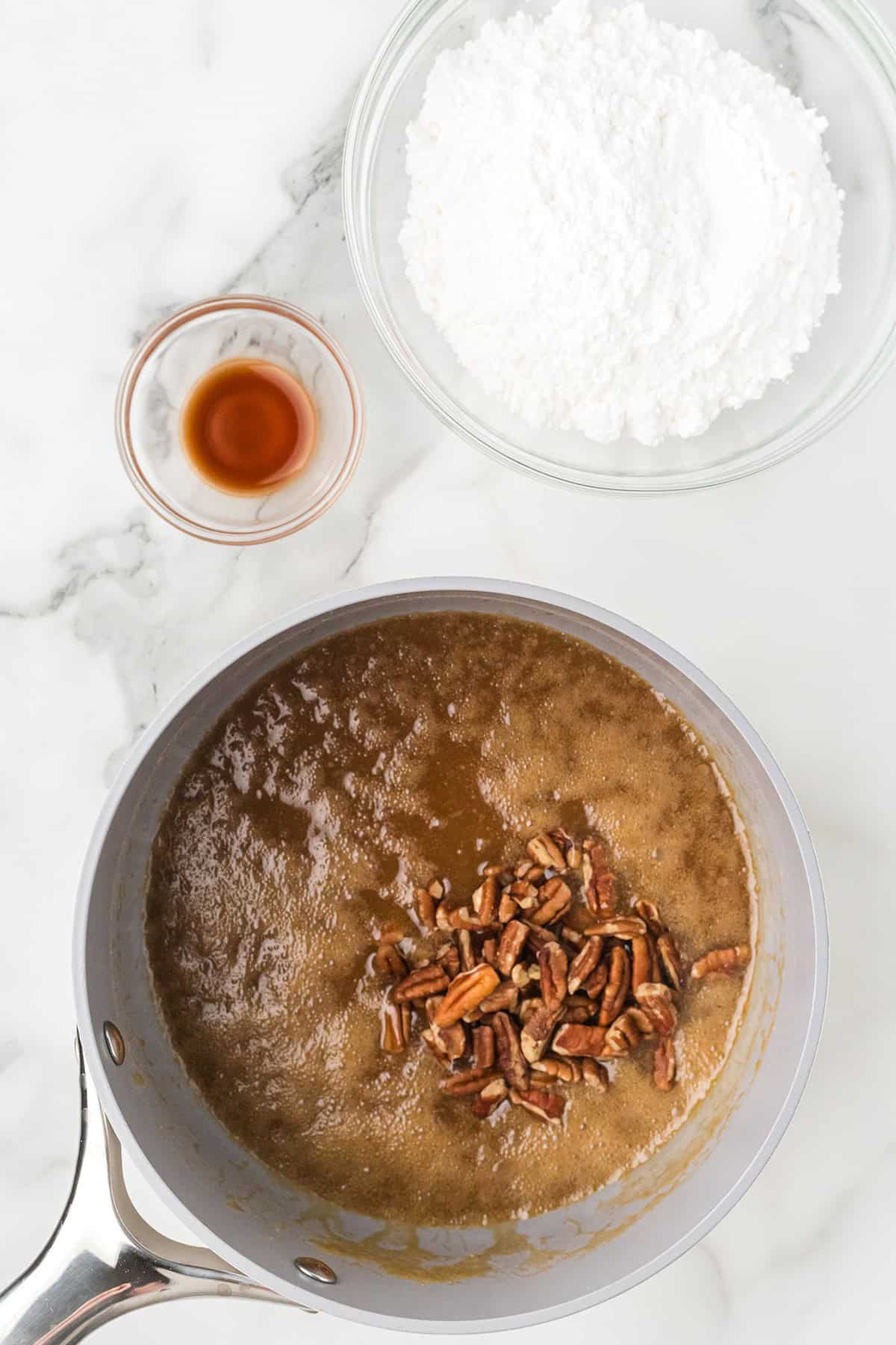 A saucepan with brown mixture and pecans next to bowls of powdered sugar and vanilla on a marble surface.