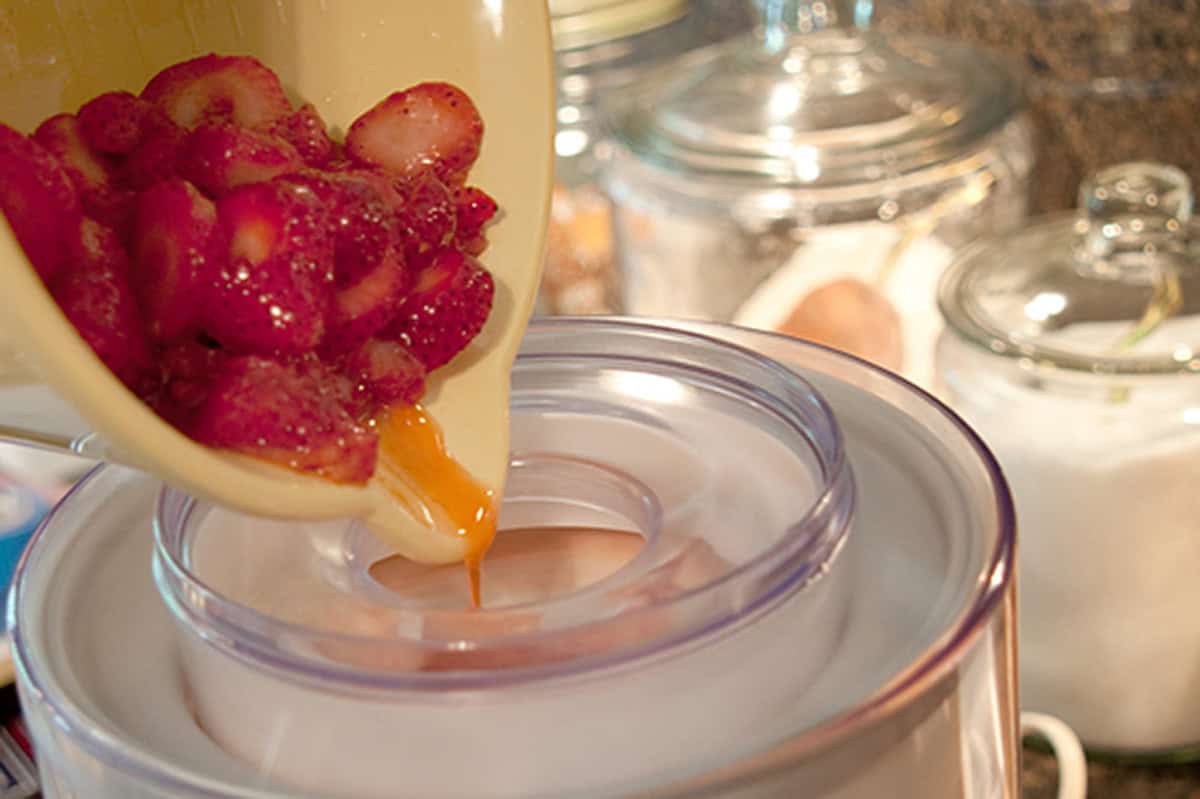 Sliced strawberries and syrup being poured into an ice cream maker in a kitchen.