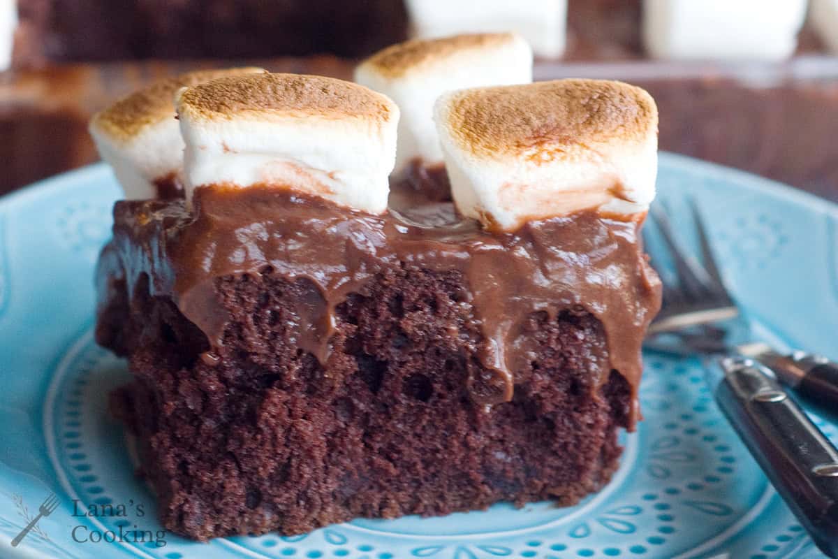 A slice of chocolate cake topped with chocolate frosting and toasted marshmallows on a blue plate with a fork and knife.