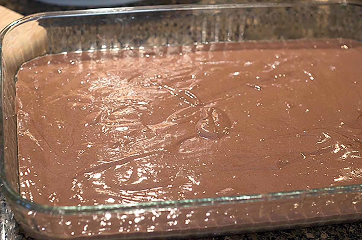 A glass baking dish filled with an even layer of uncooked chocolate cake batter on a dark countertop.