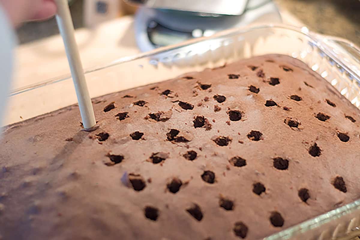 A rectangular chocolate cake in a glass baking dish with holes being poked into the surface using the handle of a wooden spoon.