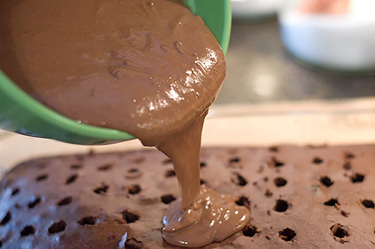Chocolate pudding is being poured from a bowl onto a chocolate cake with holes poked in the top.