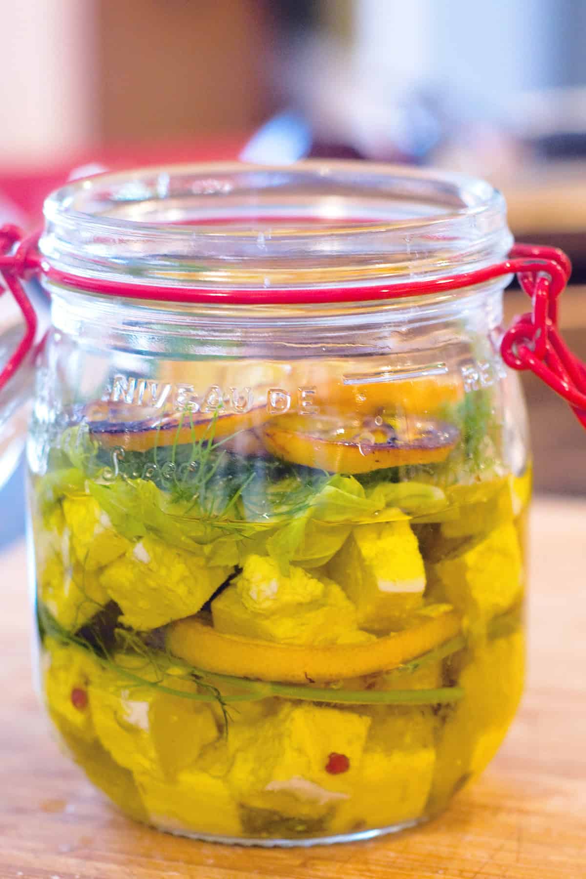A glass jar filled with marinated cheese cubes, fresh herbs, and lemon slices, submerged in oil, placed on a wooden surface.