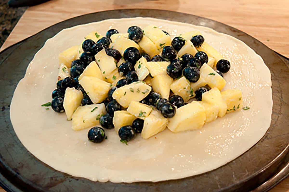 Unbaked pie crust topped with chopped pineapple, blueberries, and herbs, placed on a round baking sheet.
