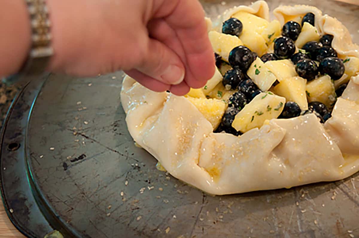 A hand sprinkles sugar over an unbaked rustic tart filled with blueberries and chopped apples on a baking sheet.