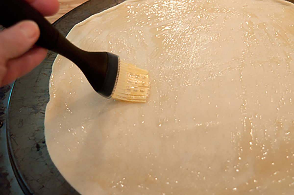 A hand using a brush to spread apple jelly onto a round sheet of dough on a dark baking tray.