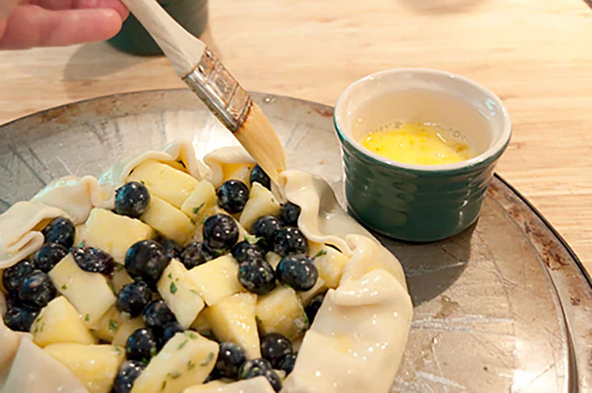 A hand uses a brush to apply egg wash to the crust of an unbaked galette filled with blueberries and diced fruit on a baking sheet. A ramekin with egg wash sits nearby.