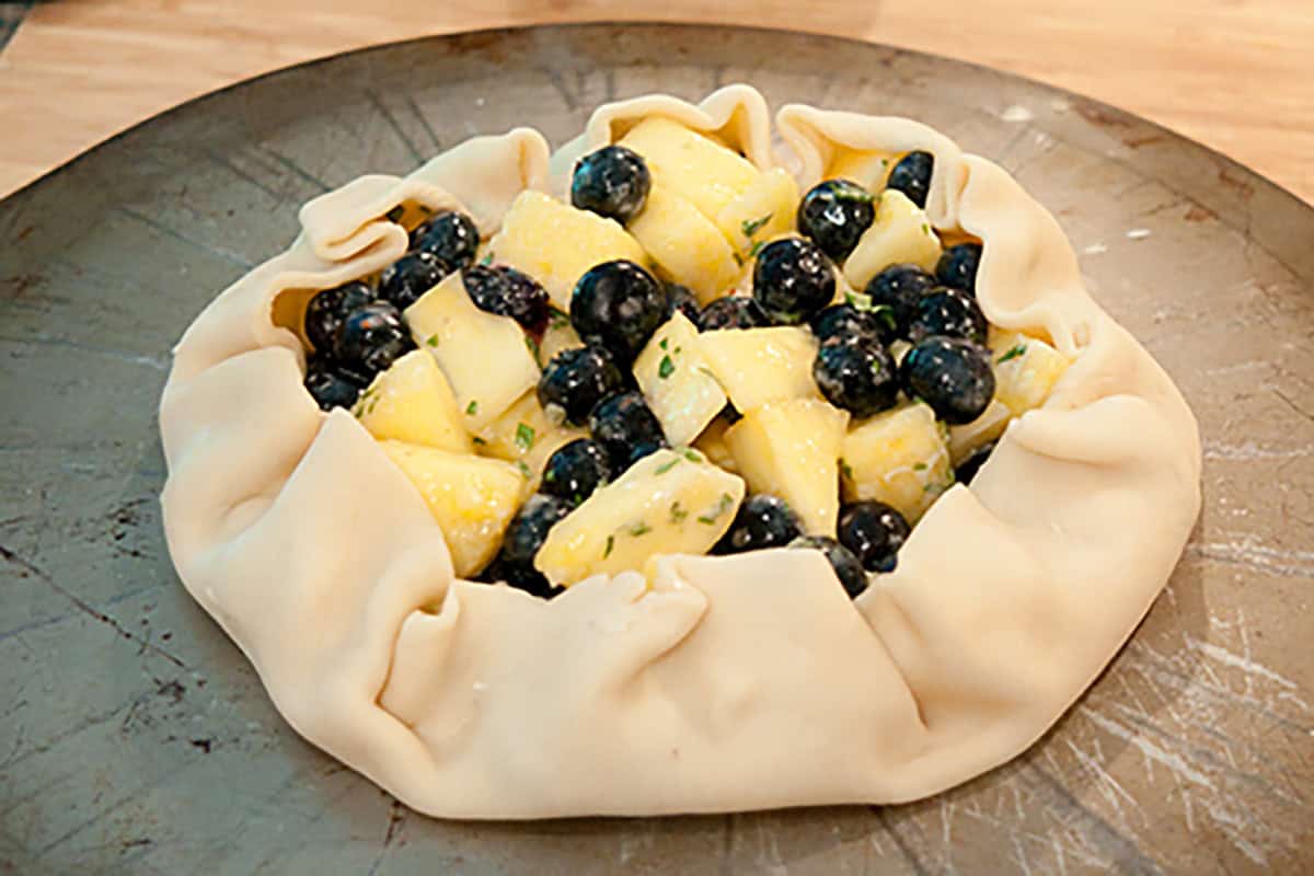 Unbaked galette with a folded pastry crust filled with chopped pineapple and whole blueberries on a baking tray.