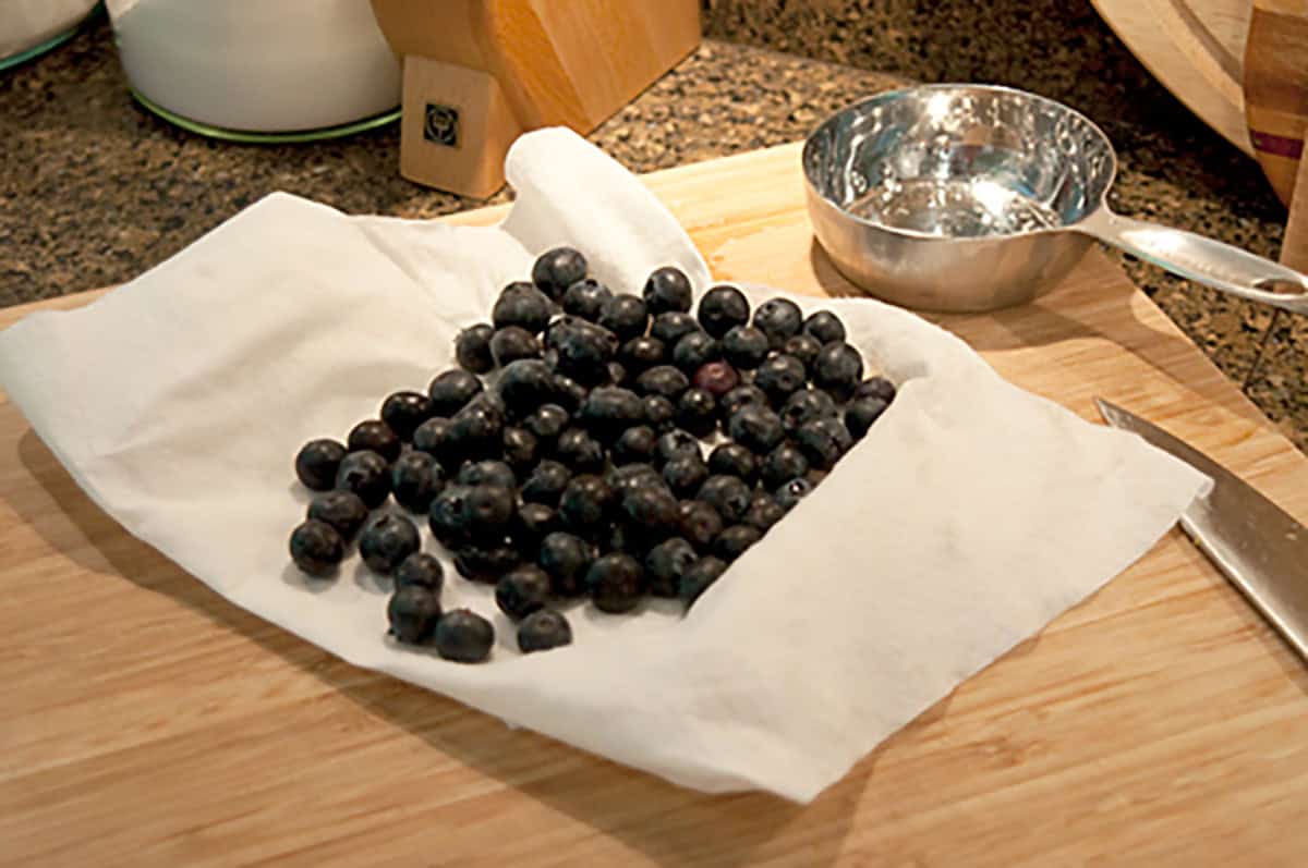A pile of fresh blueberries sits on a paper towel on a wooden cutting board, with a metal measuring cup and knife nearby.