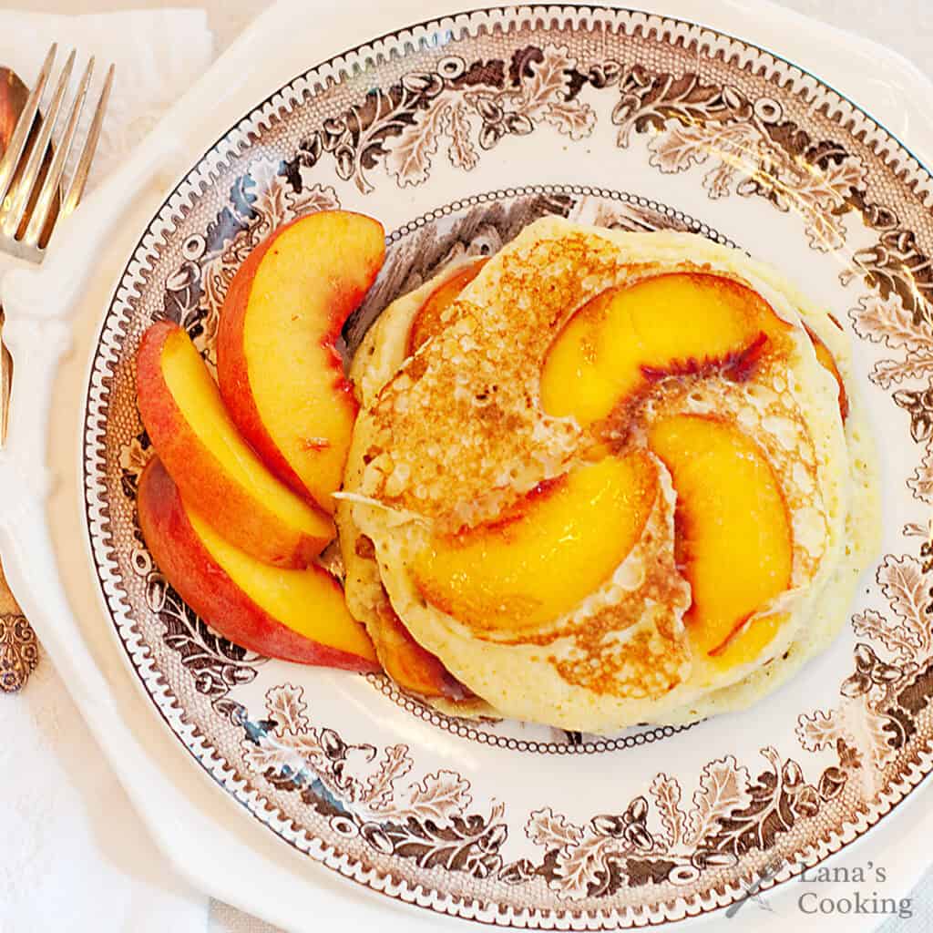 Pancakes topped with peach slices on a decorative plate, with extra peach slices on the side.