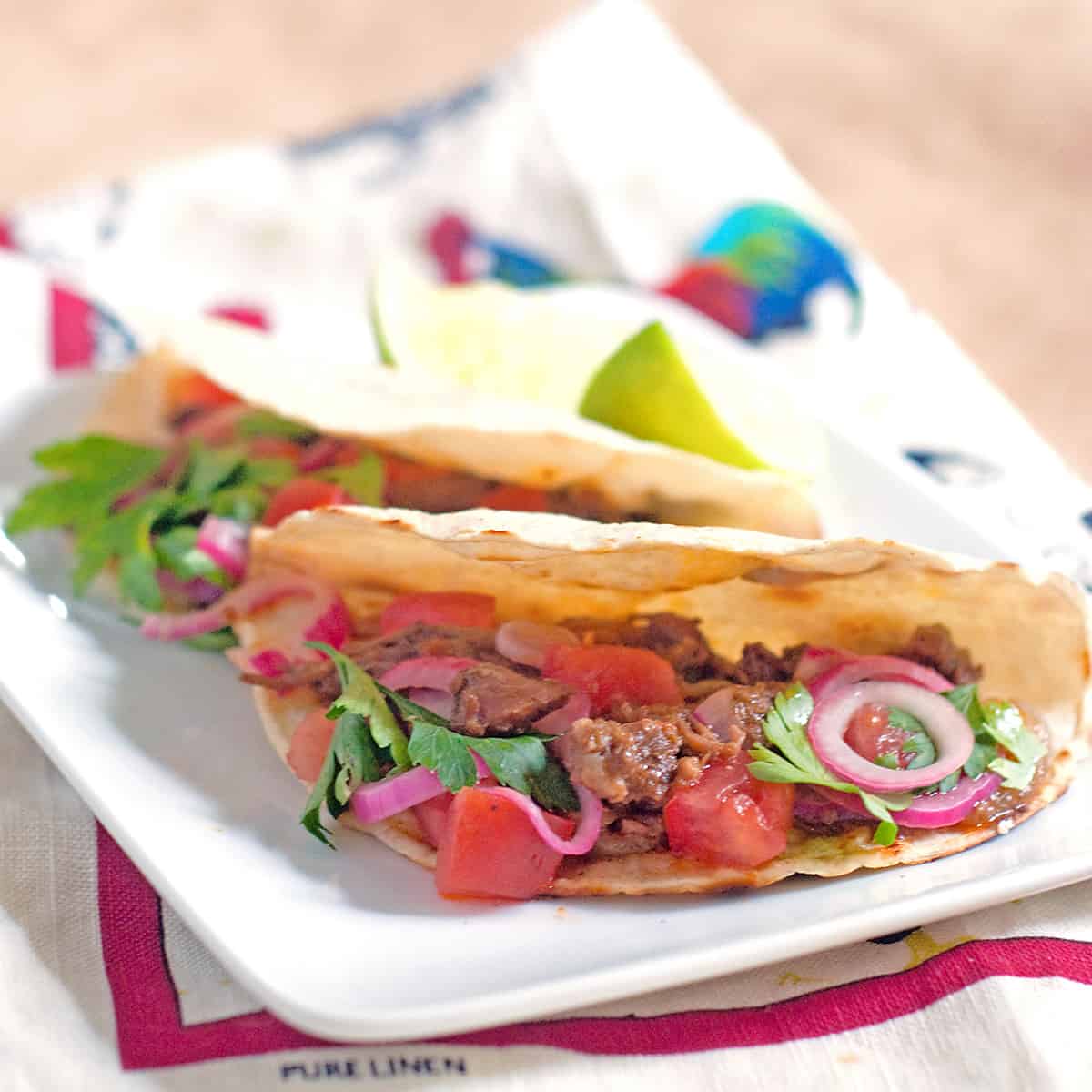 Two slow cooker shredded beef tacos on a serving plate with a kitchen towel underneath.