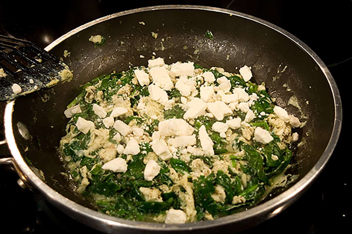 A frying pan on a stove contains scrambled eggs with spinach and is topped with crumbled feta cheese.