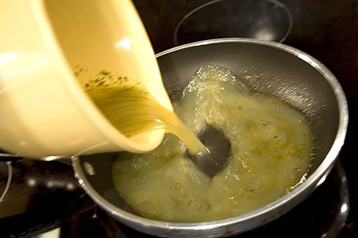 A yellow bowl pours egg whites and pesto mixture into a frying pan on a stovetop.