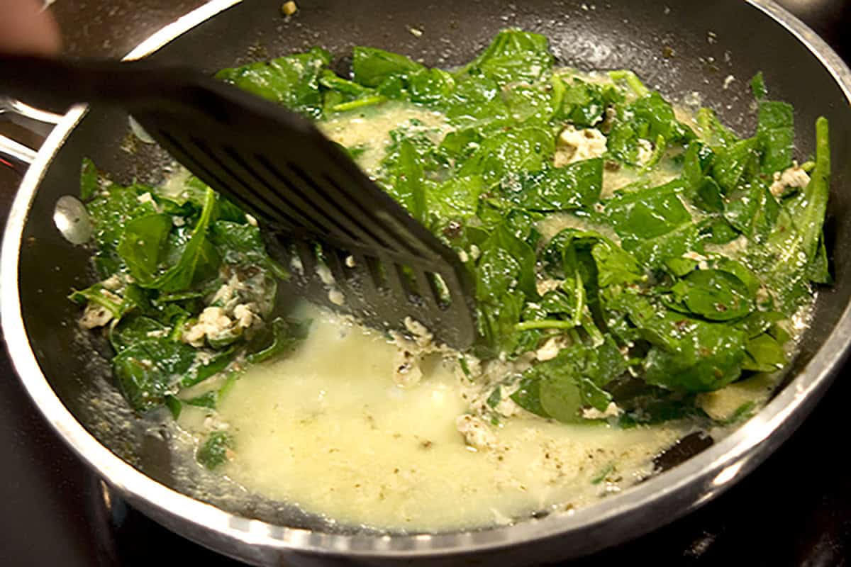 A person uses a spatula to cook spinach and egg whites in a shallow pan on a stovetop.
