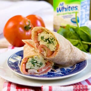 A whole wheat wrap filled with scrambled eggs, spinach, and tomato sits cut in half on a blue and white plate with fresh tomato and spinach in the background.