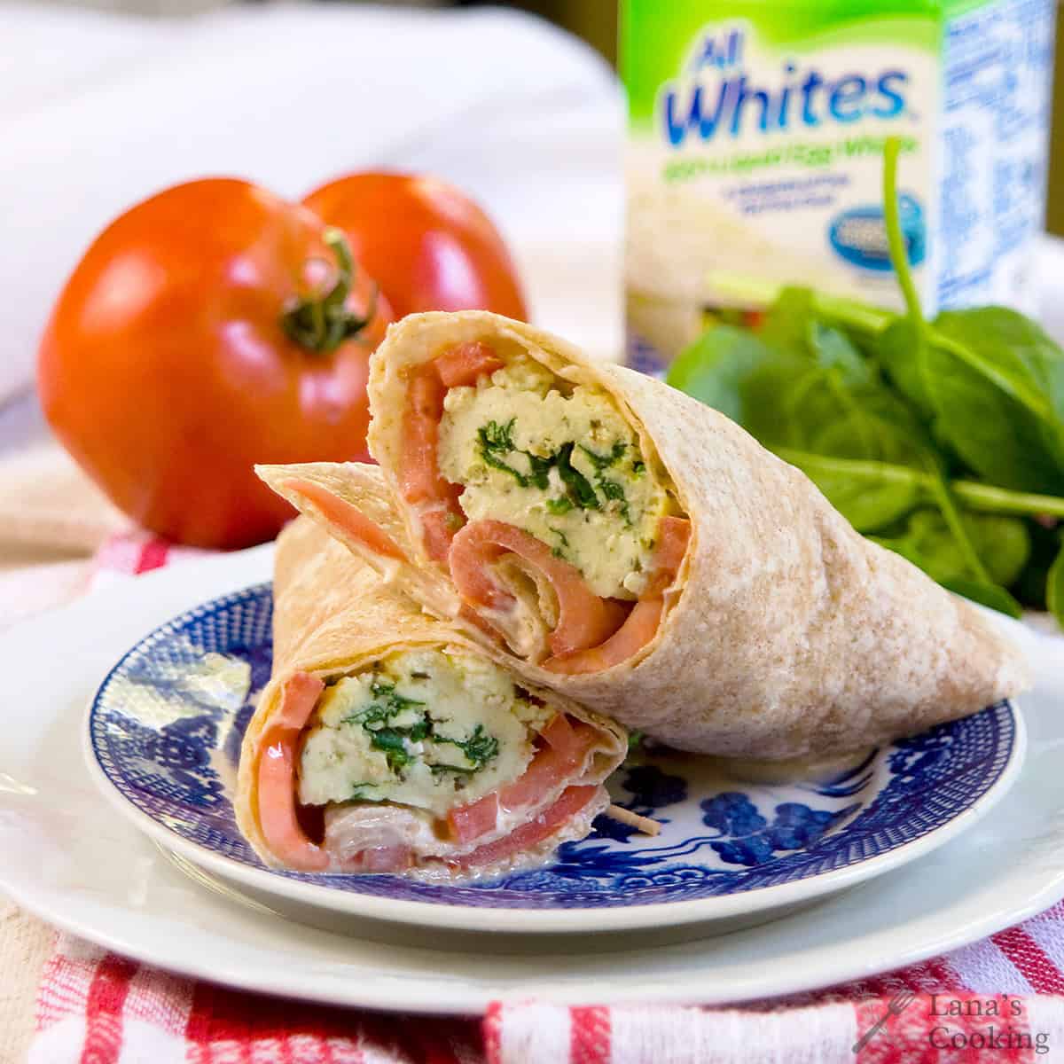 A whole wheat wrap filled with scrambled eggs, spinach, and tomato sits cut in half on a blue and white plate with fresh tomato and spinach in the background.
