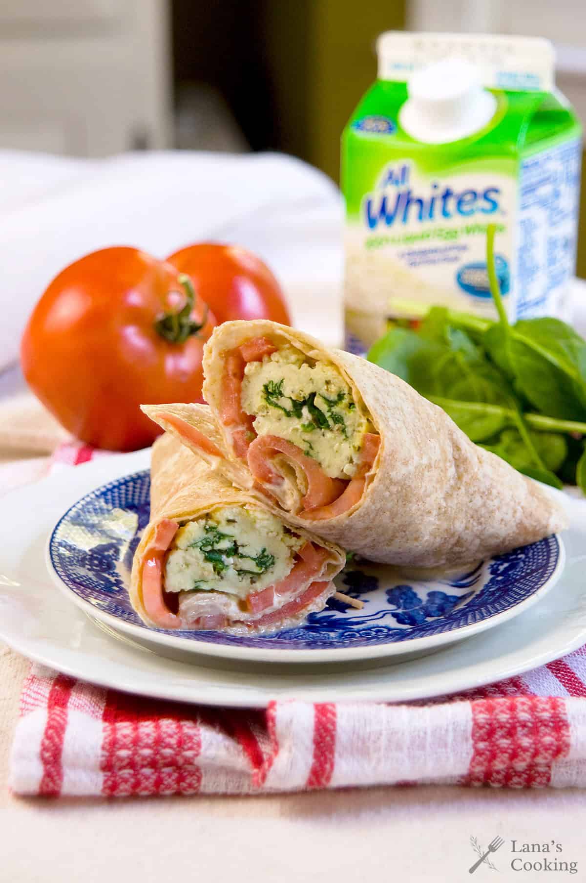 A whole wheat wrap filled with scrambled eggs, spinach, and tomato slices sits on a plate, with a tomato, spinach leaves, and a carton of egg whites in the background.