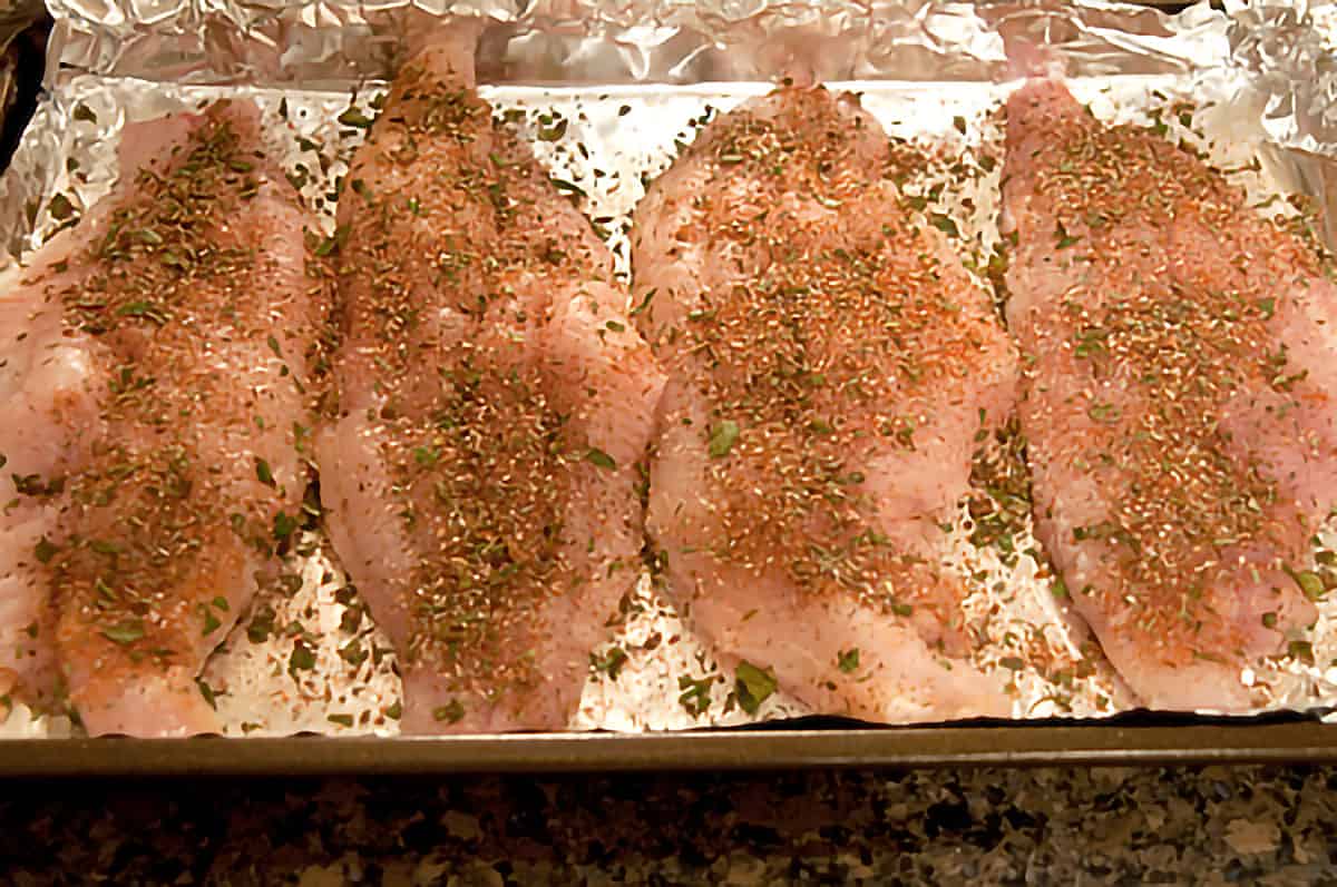 Catfish fillets on a foil-lined pan with herb and seasoning mixture sprinkled over top