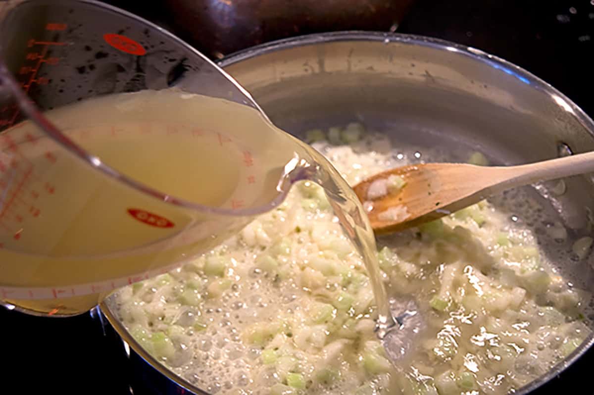 Broth is being poured from a measuring cup into a pan of saut&eacute;ing diced onions stirred with a wooden spoon.