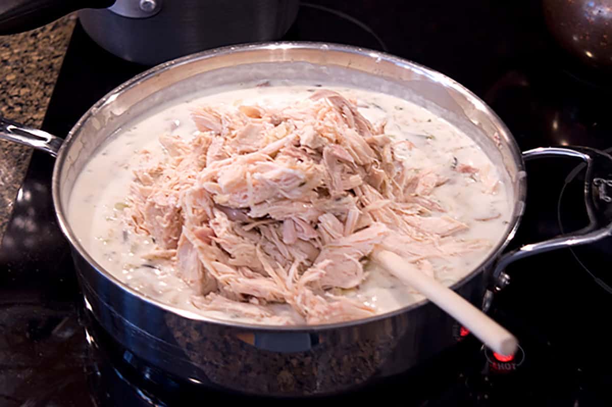 Shredded chicken in a creamy white sauce being stirred in a large pan on a stovetop.