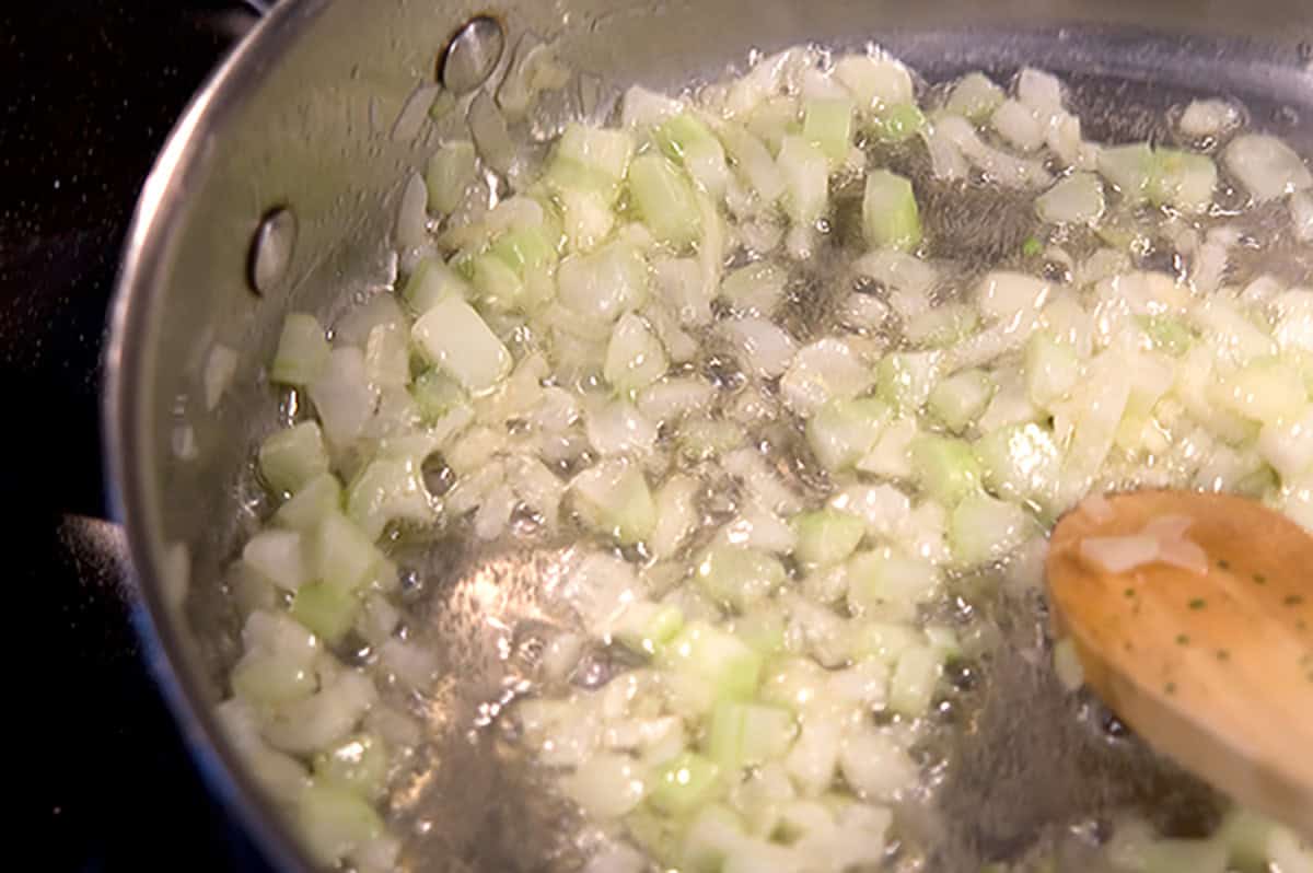 Chopped onions and celery being saut&eacute;ed in a metal pan with a wooden spoon.