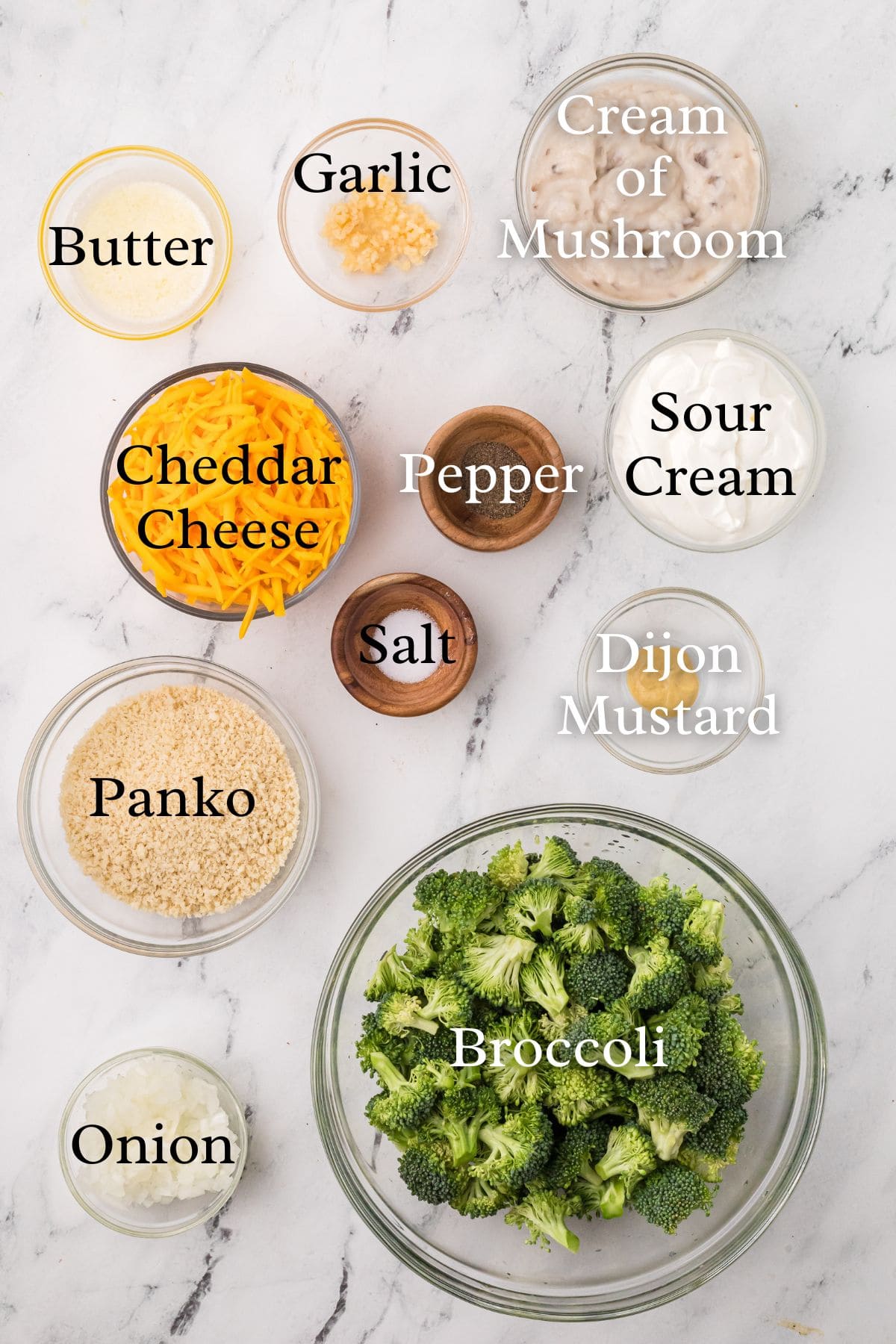 A marble surface displays labeled bowls containing butter, minced garlic, cream of mushroom soup, shredded cheddar cheese, pepper, sour cream, panko breadcrumbs, salt, Dijon mustard, chopped onion, and broccoli florets.