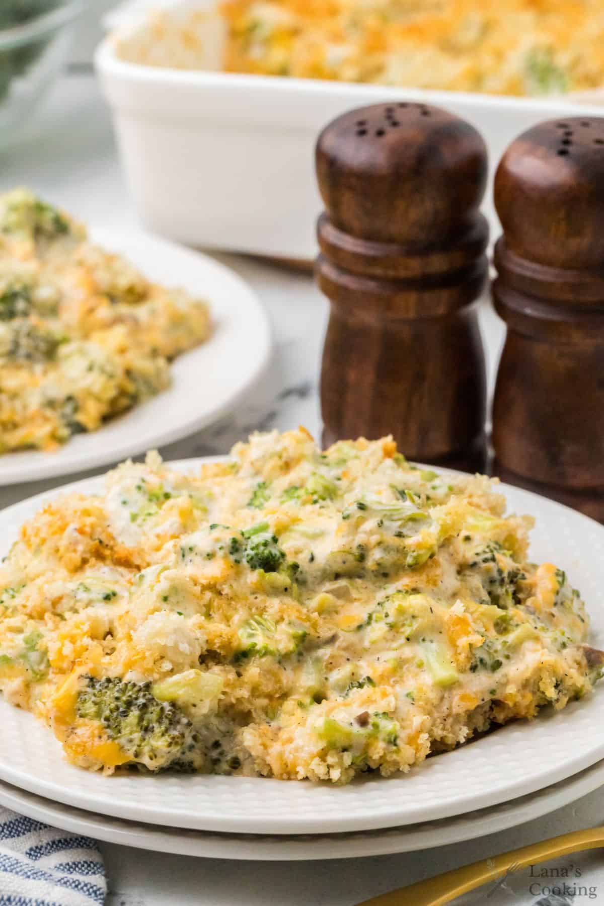 A serving of broccoli and cheese casserole on a white plate, with a larger casserole dish and two wooden salt and pepper shakers in the background. The casserole contains broccoli florets and a creamy cheese sauce.