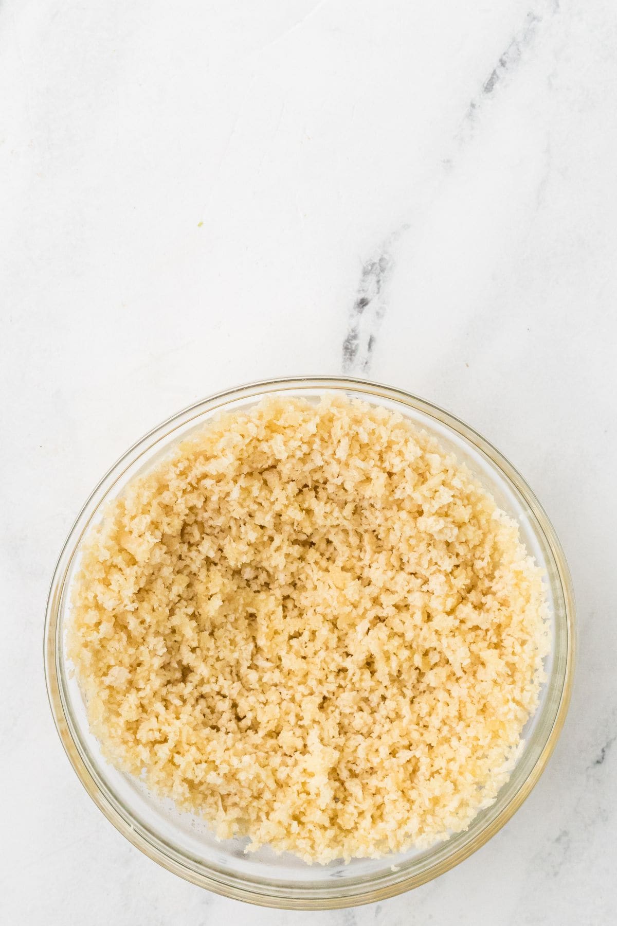 A clear glass bowl filled with finely grated cauliflower, resembling rice, sits on a white marble surface.