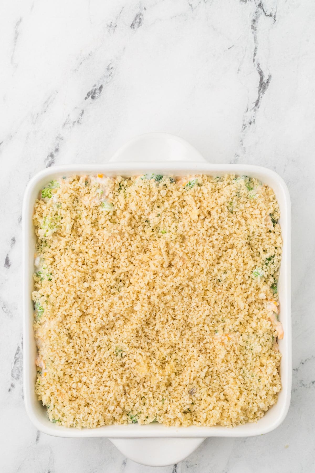A square white baking dish filled with an unbaked casserole topped with an even layer of breadcrumbs, sitting on a white marble surface.
