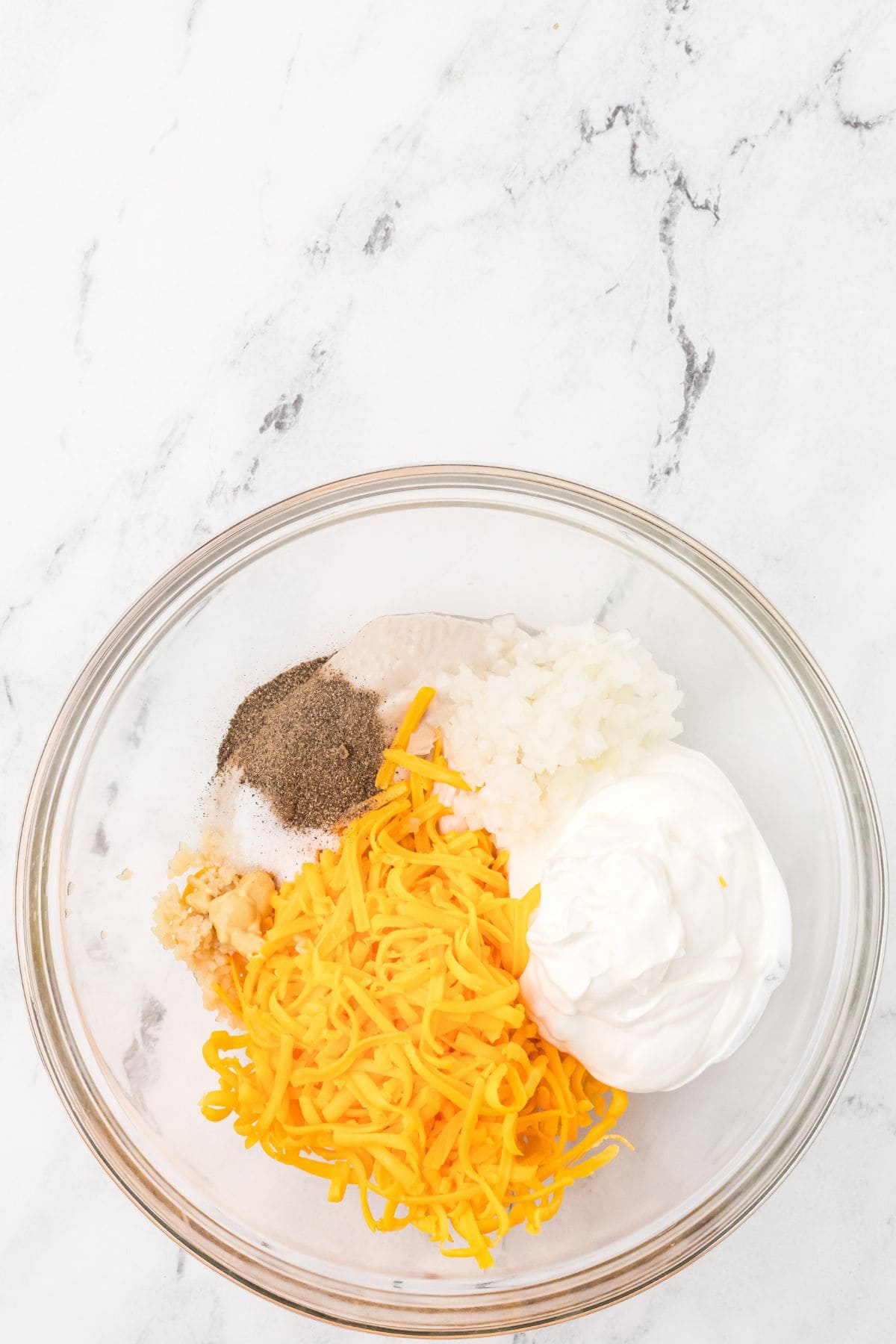 A clear glass bowl on a white marble surface containing shredded cheddar cheese, chopped onion, sour cream, minced garlic, black pepper, and salt, each ingredient arranged in separate sections.