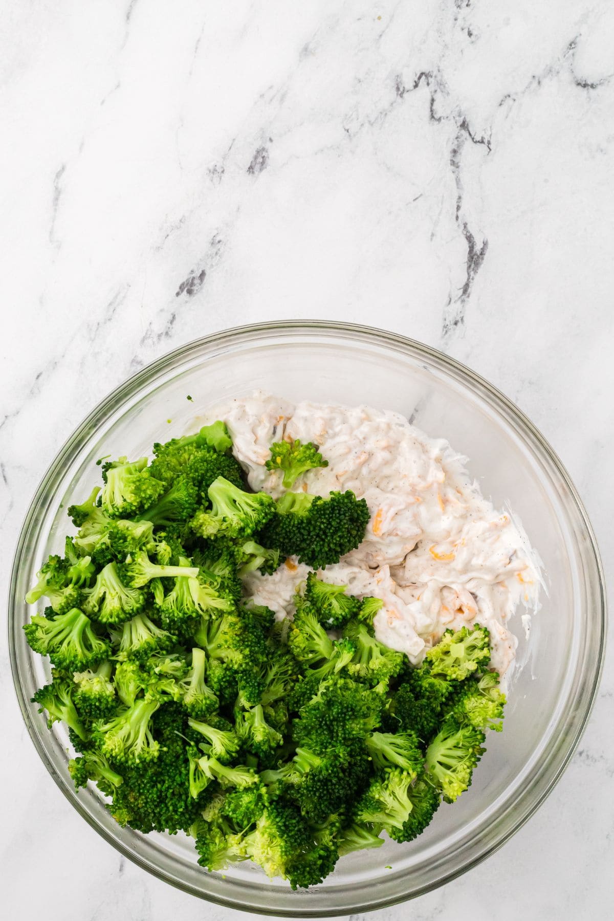 A glass bowl containing chopped broccoli on one side and a creamy white mixture on the other, set on a white marble surface.