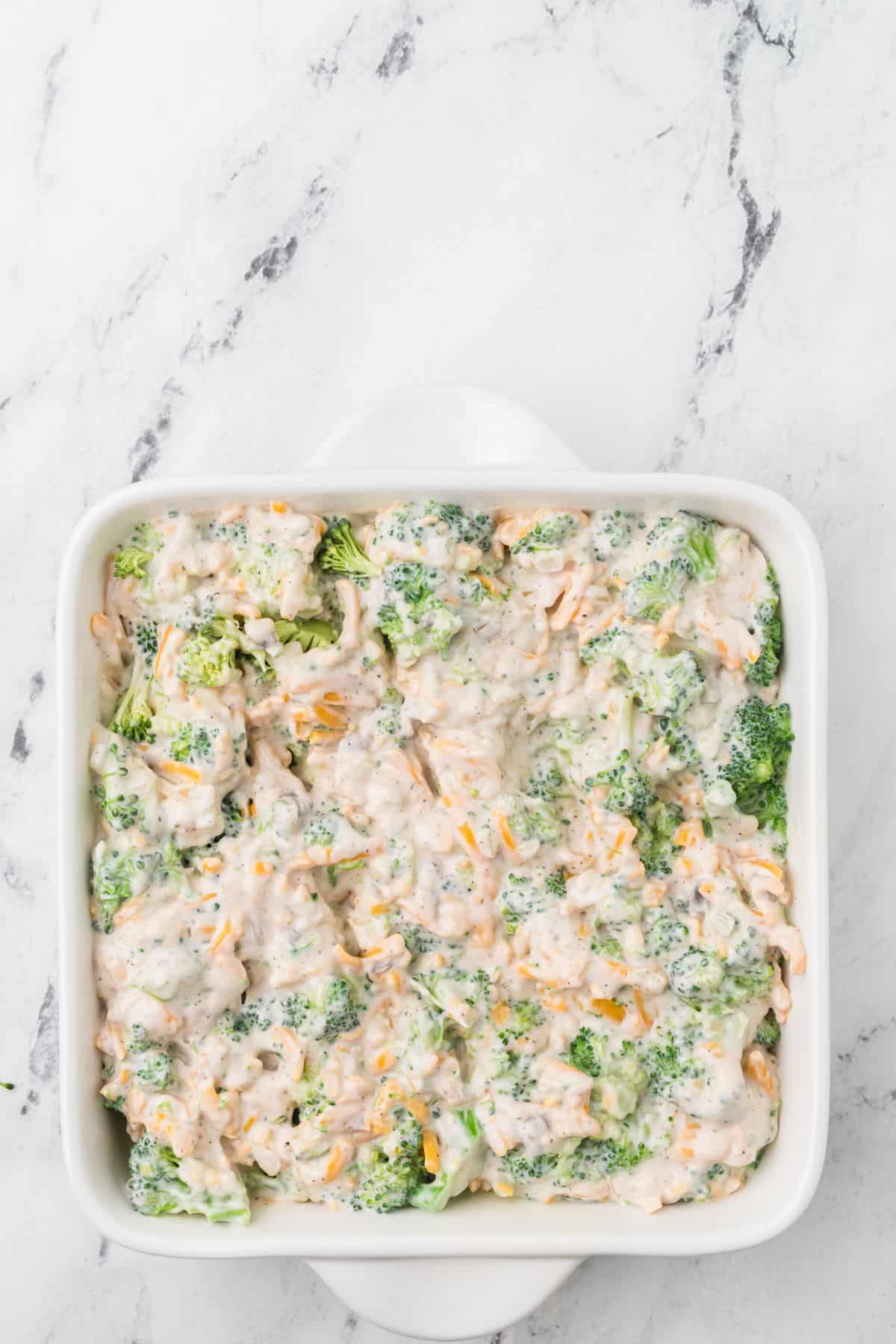 A white square baking dish filled with broccoli florets mixed with a creamy white sauce and shredded cheese, sitting on a white marble surface.
