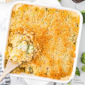 A white square baking dish filled with a casserole topped with a golden breadcrumb crust. A wooden spoon is lifting a serving, showing a creamy mixture with visible broccoli pieces underneath.