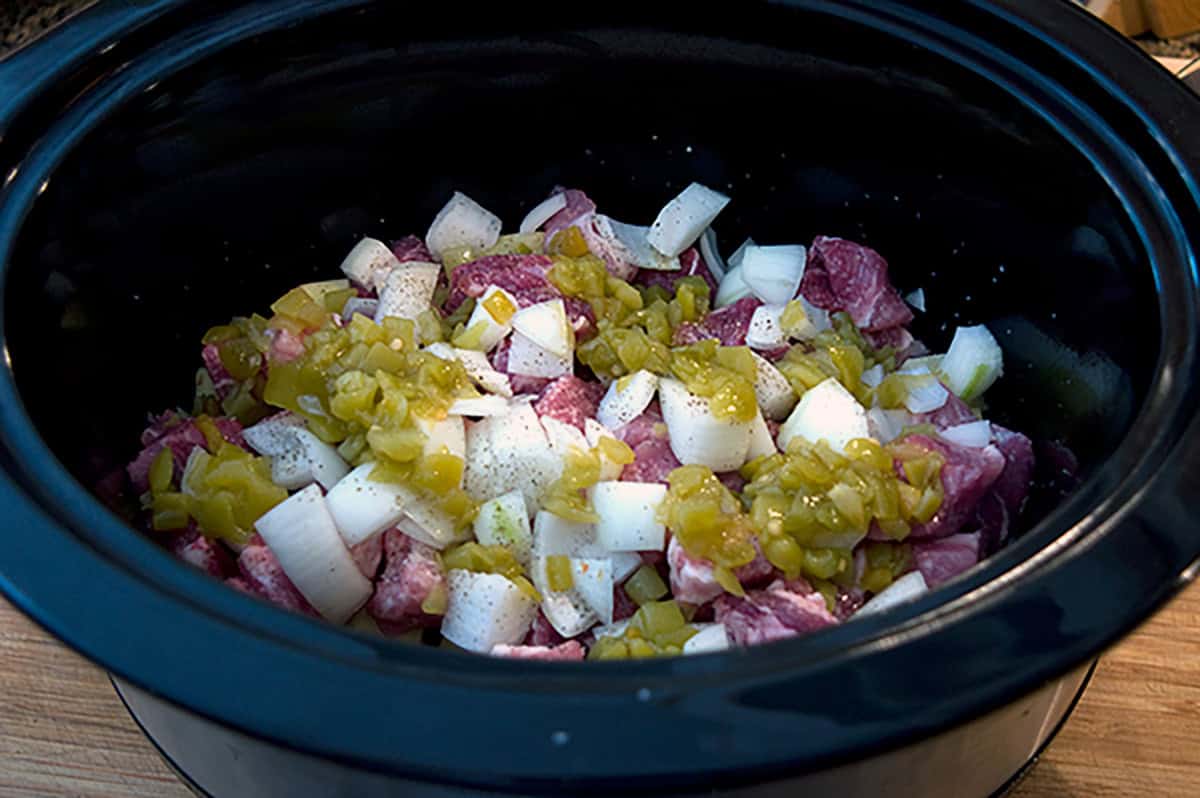 Diced pork shoulder, onions, and green chilies in a black slow cooker, ready to be cooked.