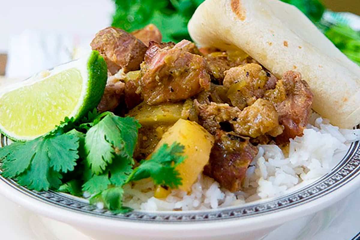 A bowl of rice topped with stewed meat, cilantro, a lime wedge, pineapple, and a folded tortilla.