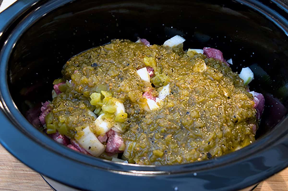 Chunks of raw meat, diced onions, and green sauce in a black slow cooker before cooking.