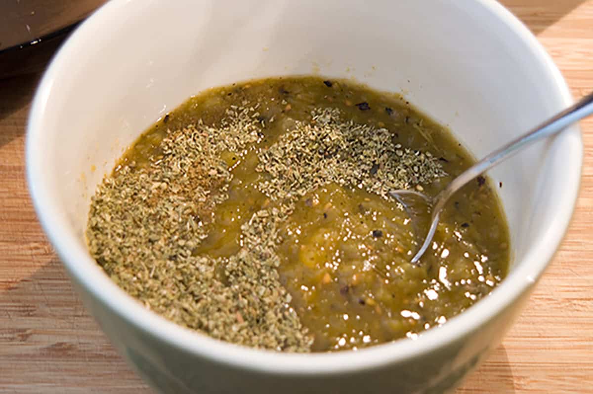 Green salsa in a white bowl, topped with dried herbs, with a spoon inside the bowl.
