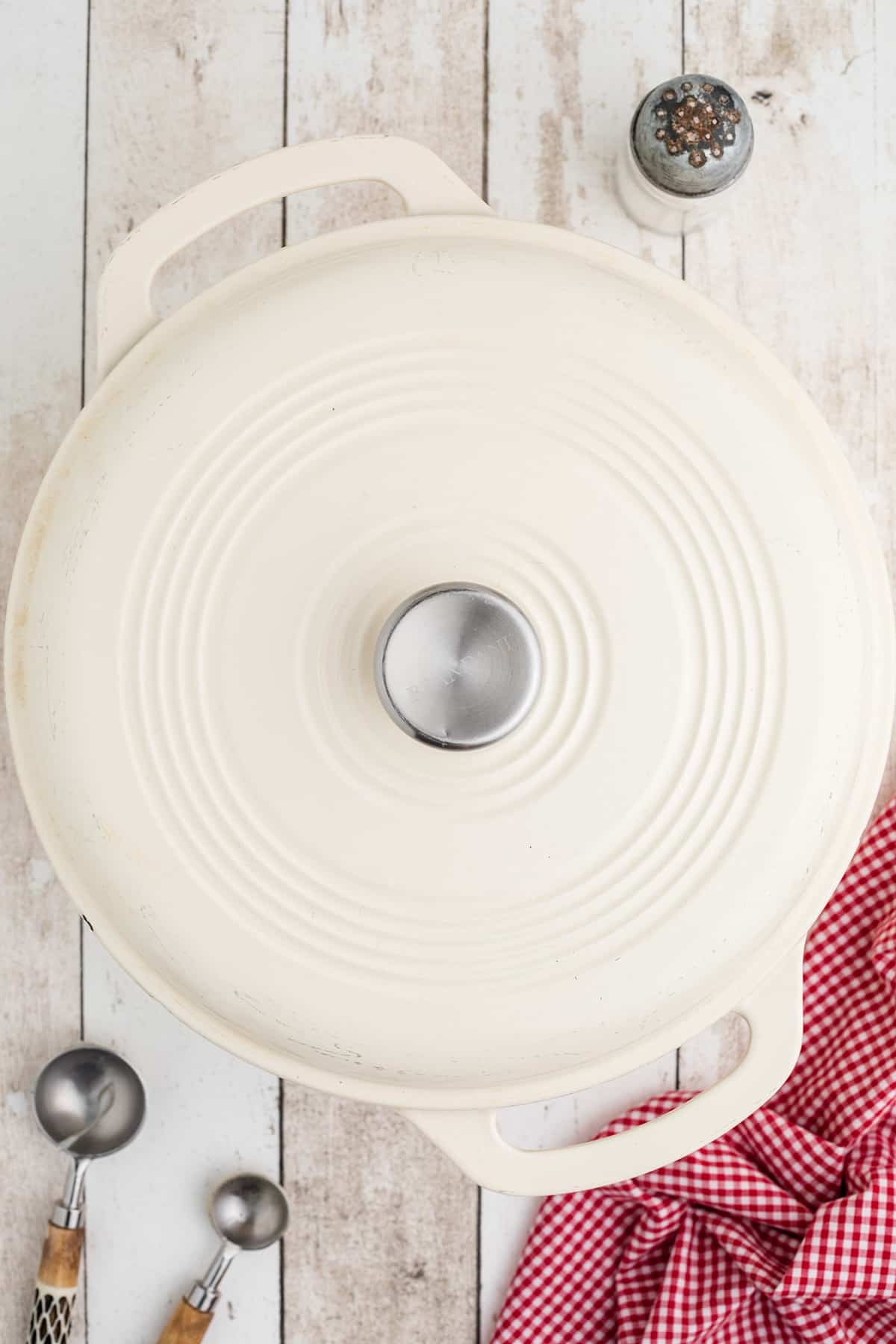 White Dutch oven with lid on, pepper shaker, measuring spoons, and red checkered cloth on wooden surface.