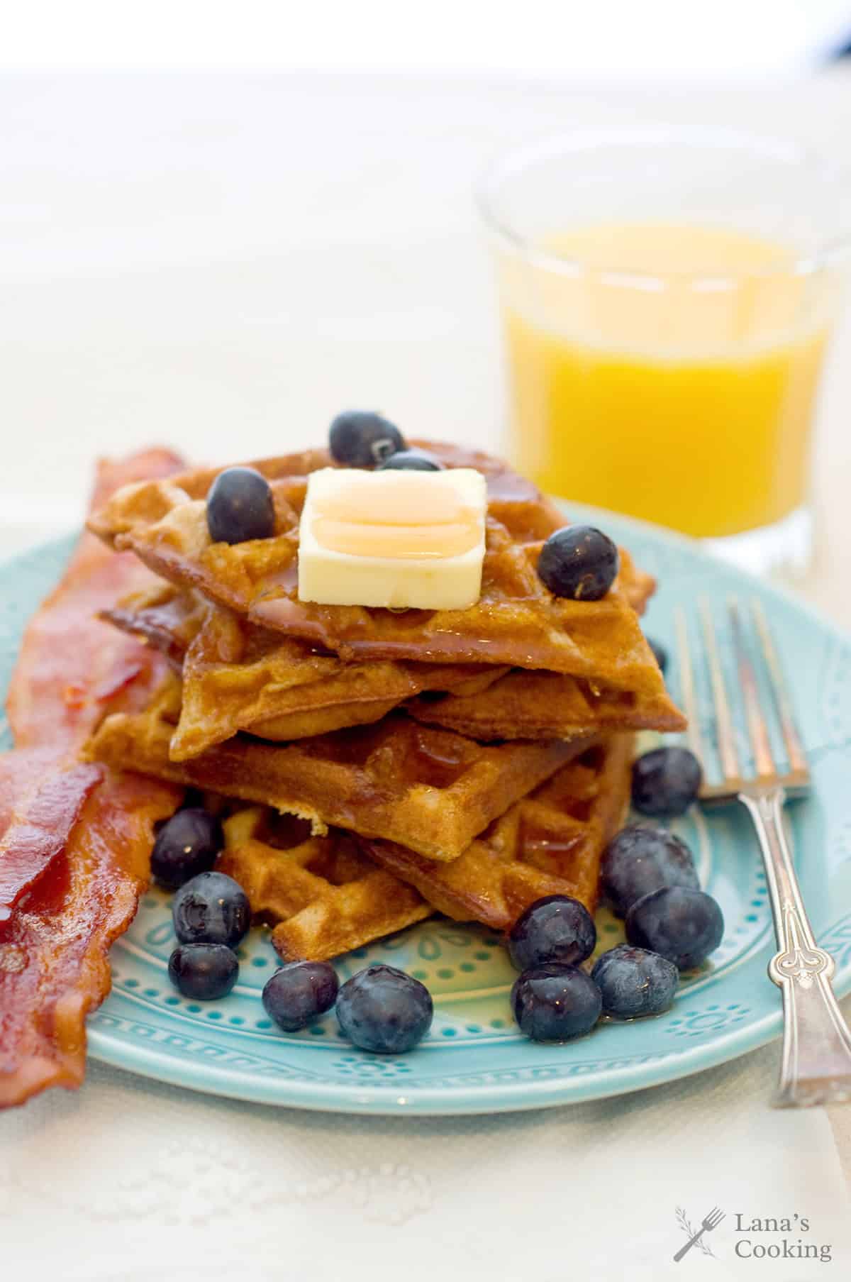 A plate of waffles with butter, blueberries, bacon, and a glass of orange juice on the side.