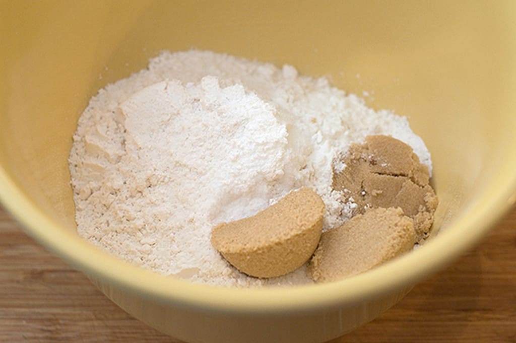 A yellow bowl with brown sugar and flour on a wooden surface.