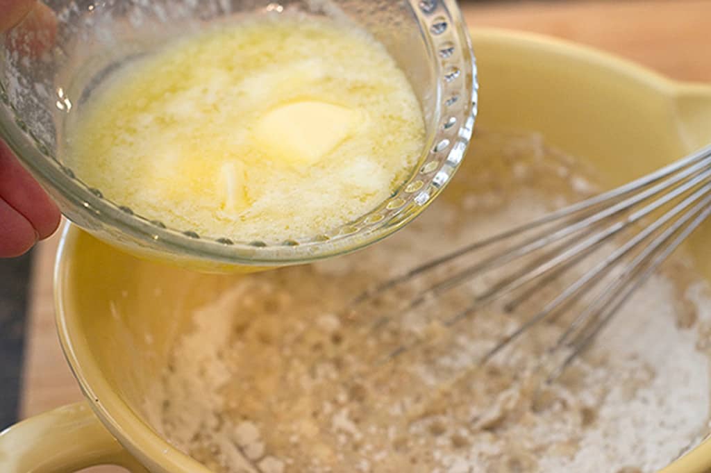 A hand pours melted butter from a glass bowl into a mixing bowl with batter and a metal whisk.
