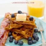 Stack of waffles with butter and blueberries, served with bacon and orange juice on a blue plate.